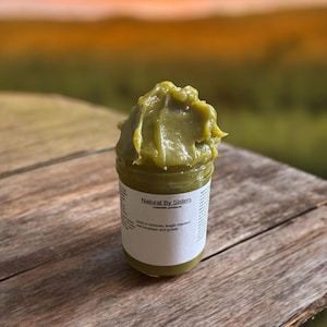 Pode incluir: Um frasco de produto cosm&eacute;tico verde, rotulado "Natural By Sisters". O produto &eacute; uma pasta espessa e texturizada. O frasco est&aacute; sobre uma superf&iacute;cie de madeira, com um p&ocirc;r do sol desfocado ao fundo. O r&oacute;tulo menciona hidrata&ccedil;&atilde;o, reten&ccedil;&atilde;o de comprimento e quebra e crescimento.