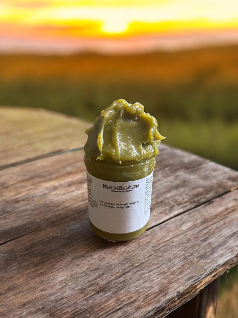 Pode incluir: Um frasco de produto cosm&eacute;tico verde, rotulado "Natural By Sisters". O produto &eacute; uma pasta espessa e texturizada. O frasco est&aacute; sobre uma superf&iacute;cie de madeira, com um p&ocirc;r do sol desfocado ao fundo. O r&oacute;tulo menciona hidrata&ccedil;&atilde;o, reten&ccedil;&atilde;o de comprimento e quebra e crescimento.