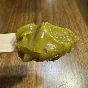 Pode incluir: Um close-up de um palito de madeira segurando uma por&ccedil;&atilde;o de subst&acirc;ncia amarelo-esverdeada. A subst&acirc;ncia tem uma apar&ecirc;ncia espessa e texturizada com manchas vis&iacute;veis. O fundo &eacute; uma superf&iacute;cie de madeira com um tom marrom quente.