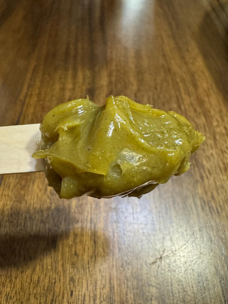 Pode incluir: Um close-up de um palito de madeira segurando uma por&ccedil;&atilde;o de subst&acirc;ncia amarelo-esverdeada. A subst&acirc;ncia tem uma apar&ecirc;ncia espessa e texturizada com manchas vis&iacute;veis. O fundo &eacute; uma superf&iacute;cie de madeira com um tom marrom quente.