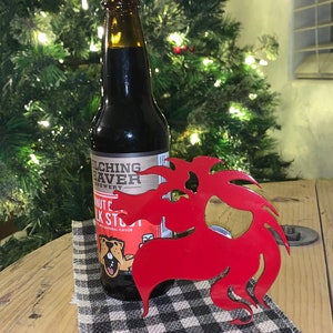 Może przedstawiać: Czerwony metalowy otwieracz do butelek w kształcie kreskówkowego bobra z butelką piwa "Welching Beaver Brewery" Peanut Butter Milk Stout z przodu. Otwieracz do butelek leży na czarno-białym wzorzystym materiale.
