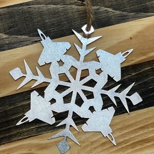 May include: A silver metal snowflake ornament with a string for hanging. The snowflake is designed with six points, each point featuring the silhouette of a fighter jet.