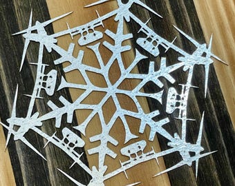 May include: A silver snowflake ornament with a detailed design, cut from paper. The snowflake is on a wooden background with dark brown and light brown stripes.