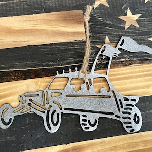 May include: A silver metal ornament shaped like a dune buggy with a flag on top. The ornament is hanging from a string against a wooden background.