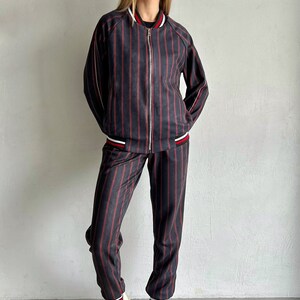 May include: A striped tracksuit with a black and red striped pattern. The jacket has a zipper closure and a white and red trim. The pants have a cuffed hem with a white and red trim. The model is wearing white sneakers.
