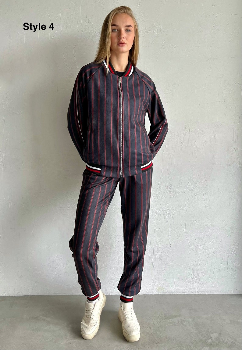 May include: A striped tracksuit with a black and red striped pattern. The jacket has a zipper closure and a white and red trim. The pants have a cuffed hem with a white and red trim. The model is wearing white sneakers.