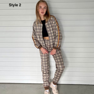 May include: A brown and beige checkered tracksuit with yellow and red stripes on the sleeves and legs. The model is wearing a black crop top underneath the jacket. The tracksuit is styled with white sneakers.