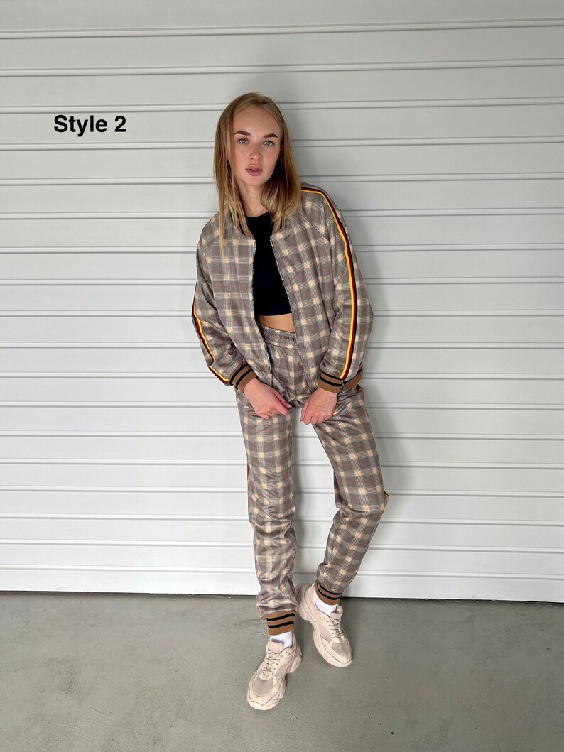 May include: A brown and beige checkered tracksuit with yellow and red stripes on the sleeves and legs. The model is wearing a black crop top underneath the jacket. The tracksuit is styled with white sneakers.