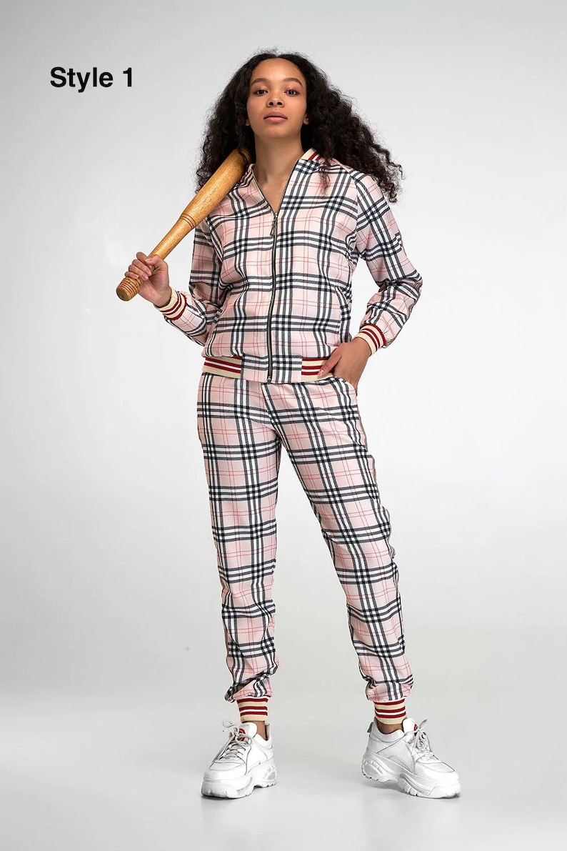 May include: A pink and black plaid tracksuit with a zippered jacket and matching pants. The jacket has a white and red stripe on the cuffs and waistband. The pants have a white and red stripe on the cuffs. The model is wearing white sneakers.