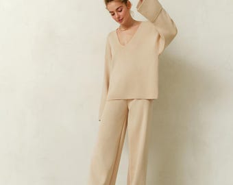 May include: A beige knitwear set consisting of a long-sleeved sweater with a V-neck and wide-leg pants. The outfit is paired with matching beige flats. The ensemble is made of a soft, comfortable material.