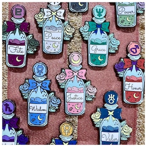 May include: Enamel pins featuring potion bottle designs with various labels like "Fate," "Peace," and "Grace." Each pin has a bow, floral accents, and a crescent moon. The color palette includes blues, pinks, and purples.