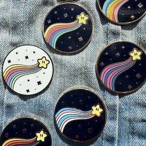 May include: A collection of round enamel pins featuring a yellow star with a rainbow trail against a dark blue background. Some pins have a white background with a rainbow trail. The pins have a gold border and are scattered on a denim surface.