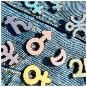 Puede incluir: Una colección de broches de esmalte en varios colores pastel, incluyendo azul claro, rosa, amarillo y verde, con contornos dorados. Los broches presentan símbolos astrológicos y celestiales, sobre un fondo de mezclilla azul.