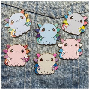 May include: Five enamel pins shaped like axolotls in various colors including light blue, pale pink, and white. Each axolotl features large black eyes, a small mouth, and colorful external gills in shades of orange, purple, blue, and pink.