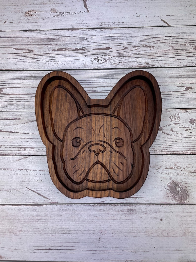 May include: A wooden tray shaped like a French Bulldog's face. The tray is dark brown and has a detailed carving of the dog's features.