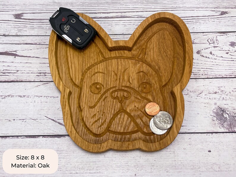 May include: A wooden tray shaped like a French Bulldog's face. The tray is 8 inches by 8 inches and is made of oak. It has a natural wood finish and is perfect for holding small items like keys, coins, or jewelry.