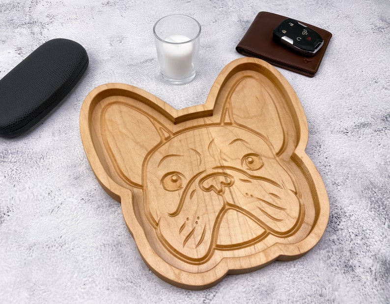May include: A wooden serving tray shaped like a French Bulldog's head. The tray is made of light brown wood and has a detailed carving of the dog's face.