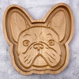 May include: A wooden serving tray shaped like a French Bulldog's face. The tray is made of light brown wood and has a detailed carving of the dog's face.