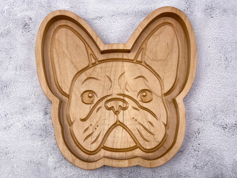 May include: A wooden serving tray shaped like a French Bulldog's face. The tray is made of light brown wood and has a detailed carving of the dog's face.