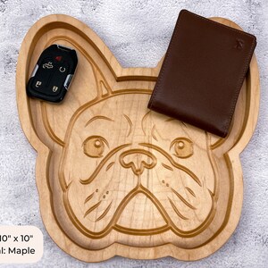 May include: A wooden tray shaped like a French Bulldog's face. The tray is made of maple wood and measures 10 inches by 10 inches. A brown leather wallet and a car key are sitting on the tray.