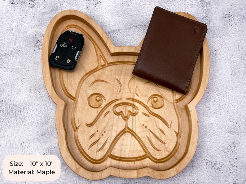 May include: A wooden tray shaped like a French Bulldog's face. The tray is made of maple wood and measures 10 inches by 10 inches. A brown leather wallet and a car key are sitting on the tray.