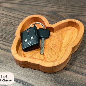 May include: A wooden key dish shaped like a French Bulldog. The dish is made of cherry wood and measures 6 inches by 6 inches. The dish is shown with a car key and a key fob inside.