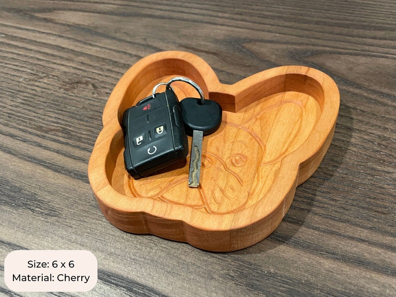 May include: A wooden key dish shaped like a French Bulldog. The dish is made of cherry wood and measures 6 inches by 6 inches. The dish is shown with a car key and a key fob inside.