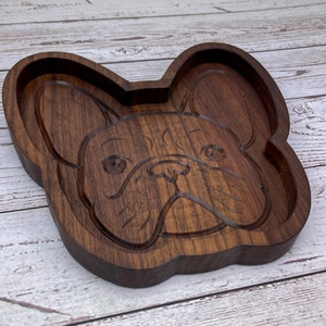 May include: A wooden tray shaped like a French Bulldog's face. The tray is made of dark brown wood and has a smooth, polished finish.