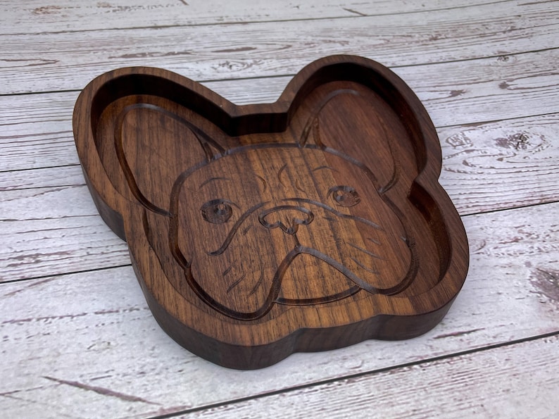 May include: A wooden tray shaped like a French Bulldog's face. The tray is made of dark brown wood and has a smooth, polished finish.