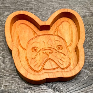 May include: A wooden dish shaped like a French Bulldog's head. The dish is carved with details of the dog's face, including its ears, nose, and eyes.