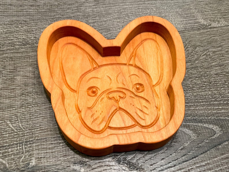 May include: A wooden dish shaped like a French Bulldog's head. The dish is carved with details of the dog's face, including its ears, nose, and eyes.