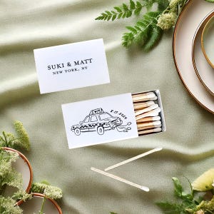 May include: White matchbox with a black and white illustration of a yellow taxi cab with the text "SUKI & MATT 8.17.2022" on the front. The matchbox is on a green tablecloth with a small bouquet of green leaves.