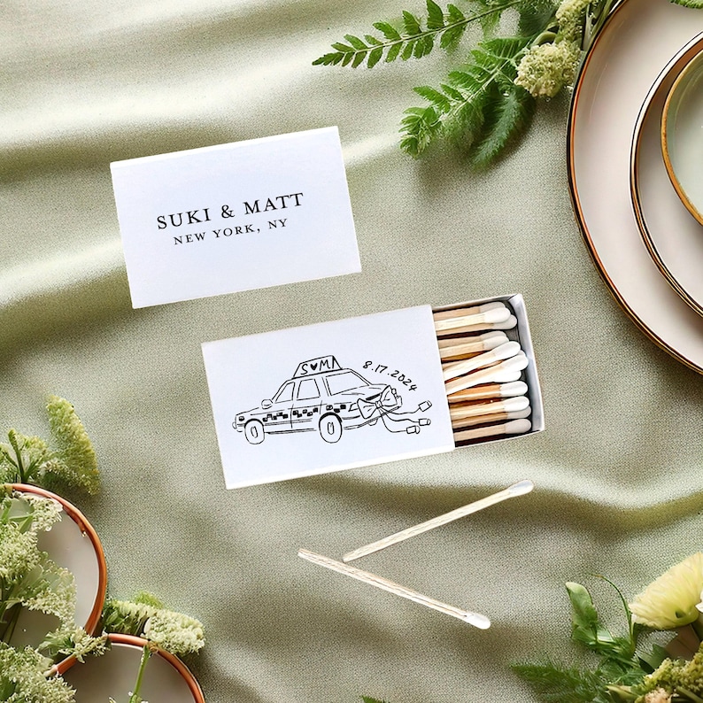 custom foil printed White matchbox with a black and white illustration of a NYC taxi cab with the personalized text "SUKI & MATT 8.17.2022" on the front. The matchbox is on a green tablecloth with a small bouquet of green leaves.