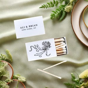 May include: White matchbox with a black illustration of two birds and a bouquet of flowers. The matchbox has a white label with the text "ALI & BRIAN NEW YORK, NY".