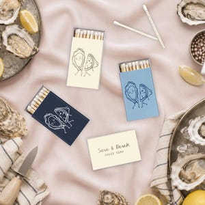 May include: Three rectangular matchboxes in cream, light blue, and navy blue, each featuring a line drawing of an oyster and a lemon wedge. One cream matchbox displays the text Sara & Derek SHUCK YEAH. The boxes contain wooden matches.