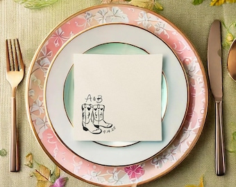 May include: A place setting with a floral-patterned plate, a white plate, and a napkin. The napkin has a black illustration of cowboy boots and the initials "A&B" with the date "9.14.25". A gold fork and knife are on either side.
