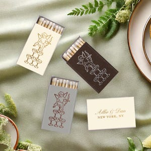 May include: Three rectangular matchboxes featuring a line drawing of a cherub sitting atop a champagne tower, available in cream, dark brown, and gray. A fourth cream-colored matchbox displays the text Allie & Dan NEW YORK, NY in gold.