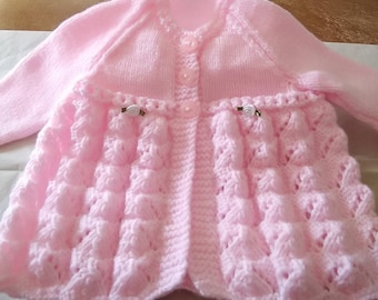 Op de afbeelding: Een roze gebreide baby-trui met knoopsluiting en een gegolfde zoom. De trui heeft een delicaat kantpatroon en is versierd met kleine witte knopen.
