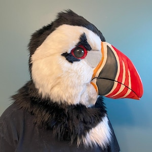 May include: A white and black furry puffin head with a red and black beak. The beak has a white stripe and a black mesh covering the eye.