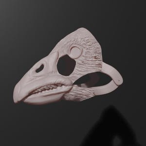 May include: A light beige dinosaur skull mask with detailed texture. The mask features a prominent beak, eye sockets, and a jawline with visible teeth. The mask is designed to be worn, with a strap visible on the side.
