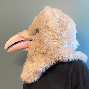 May include: A white furry bird mask with a pink beak. The mask is worn by a person with a black shirt.