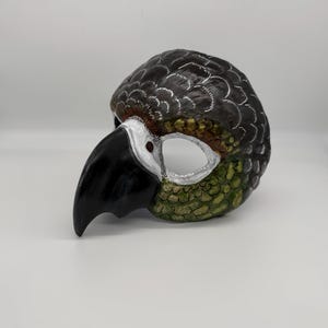 May include: A hand-painted parrot mask with a black beak and a detailed feather design. The mask features a dark gray head with white accents and green and brown detailing on the lower portion. The eye openings are white with a red dot.