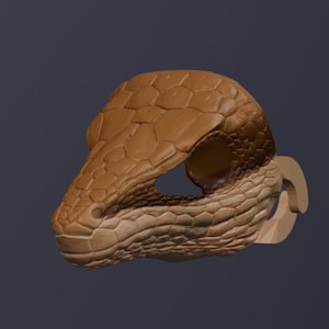 May include: A brown, reptile-themed mask with a detailed, textured surface. The mask features a snake or dragon-like head with open eye sockets and a curved, light-coloured attachment on the side. The mask is designed for costume or decorative purposes.