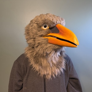 May include: A plush eagle mask with a large orange beak and yellow eyes. The mask is made of gray and brown faux fur. The person is wearing a gray zip-up hoodie.