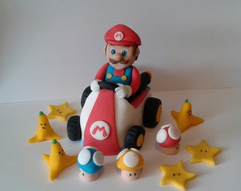 May include: A red and white Mario Kart cake topper with a figure of Mario driving a kart. The cake topper is surrounded by yellow stars, banana peels, and mushroom decorations.