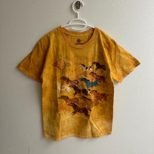 May include: Mustard yellow t-shirt with a graphic of running horses and a sun symbol. The shirt has short sleeves and a crew neck. The Mountain brand logo is visible on the collar.