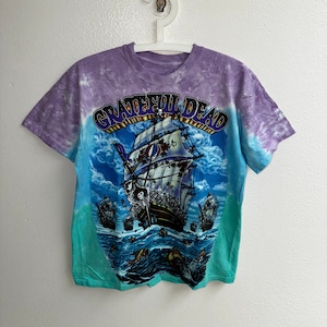 May include: A tie-dye t-shirt with a purple and green color scheme. The shirt features a graphic of a ship with skeletons on board and the text "Grateful Dead" in a yellow banner.