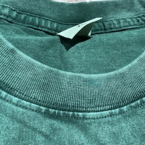 May include: Close-up of the neckline of a dark green t-shirt. The shirt has a ribbed texture and a sewn-in label.