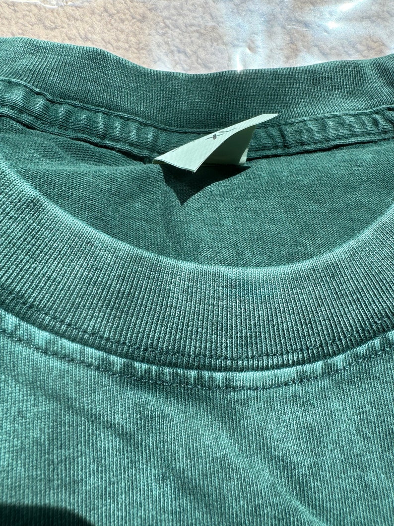 May include: Close-up of the neckline of a dark green t-shirt. The shirt has a ribbed texture and a sewn-in label.