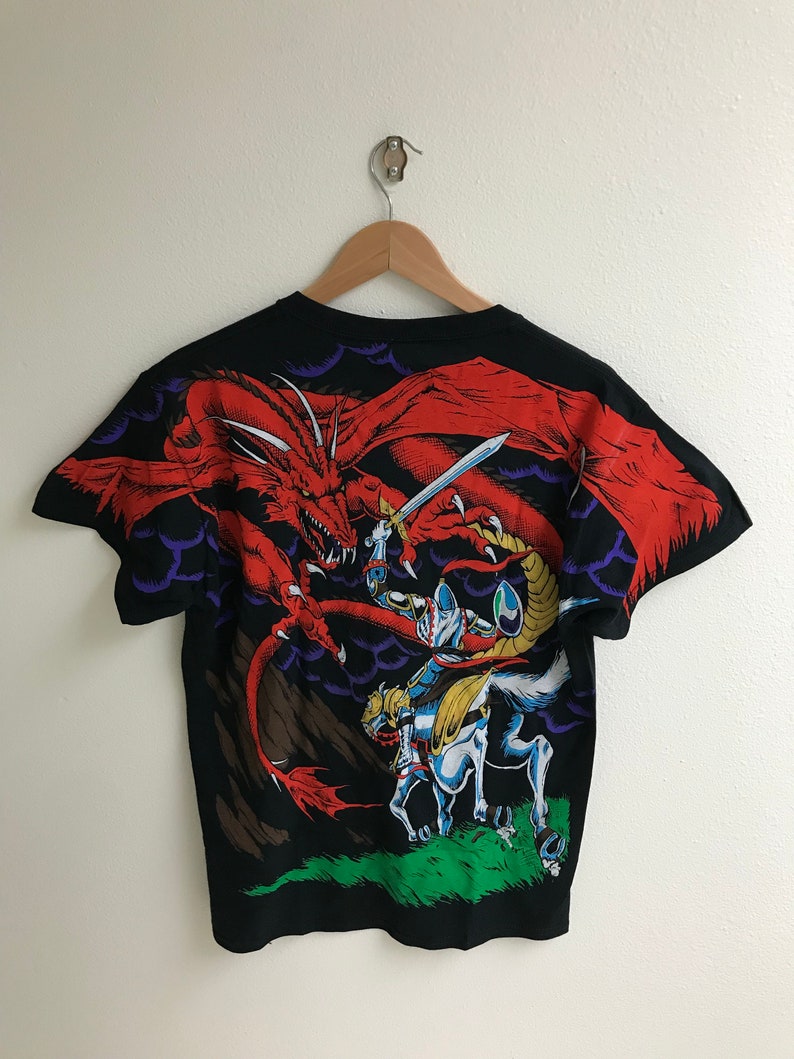 May include: Black t-shirt with a graphic print of a knight on horseback fighting a red dragon. The dragon is flying in the air and the knight is holding a sword.